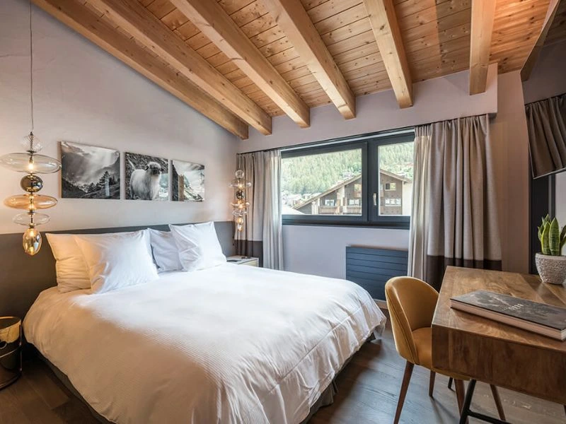 Modernes Schlafzimmer mit Holzbalkendecke, Doppelbett und Schreibtisch in alpiner Umgebung.