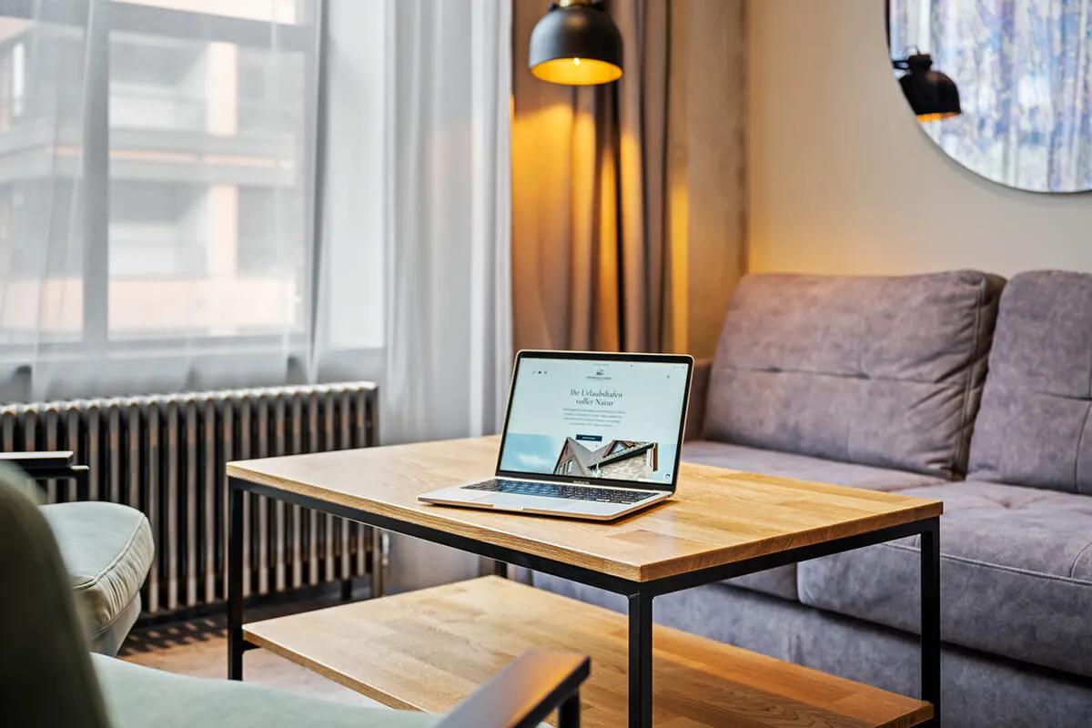 Laptop auf Wohnzimmertisch zeigt die Website einer Ferienunterkunft mit Hausfoto.