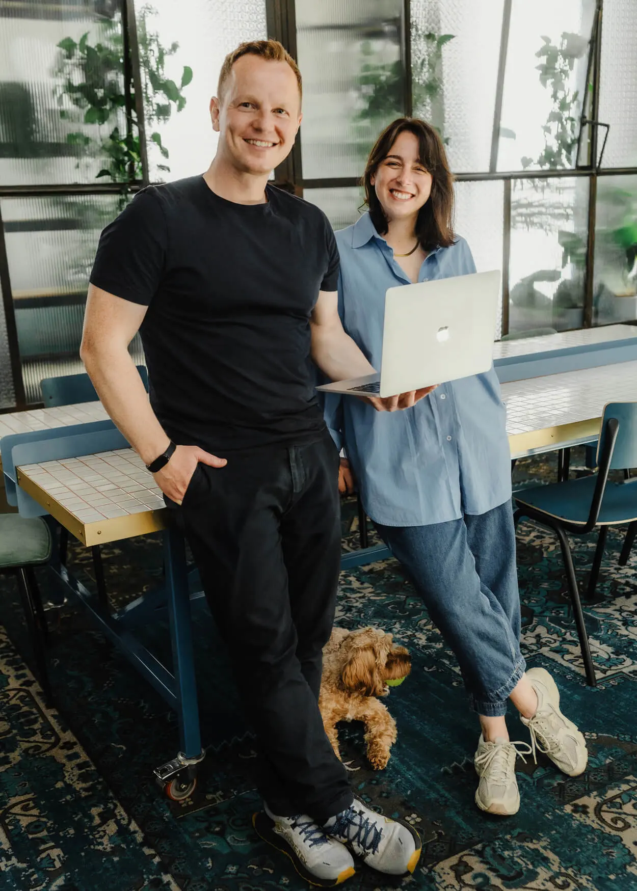 acek und Agnes stehen lächelnd mit ihrem Cavapoo Maggie im Büro.