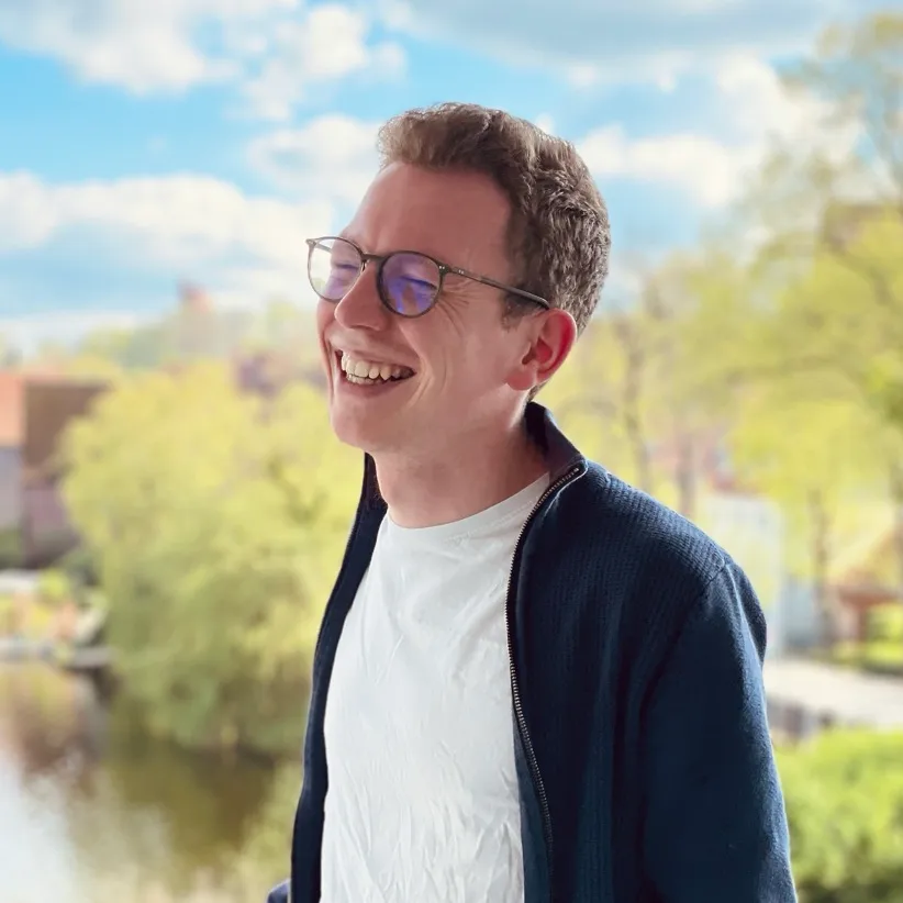 Mika steht lächelnd mit Brille vor einem hellen, grünen Außenbereich.